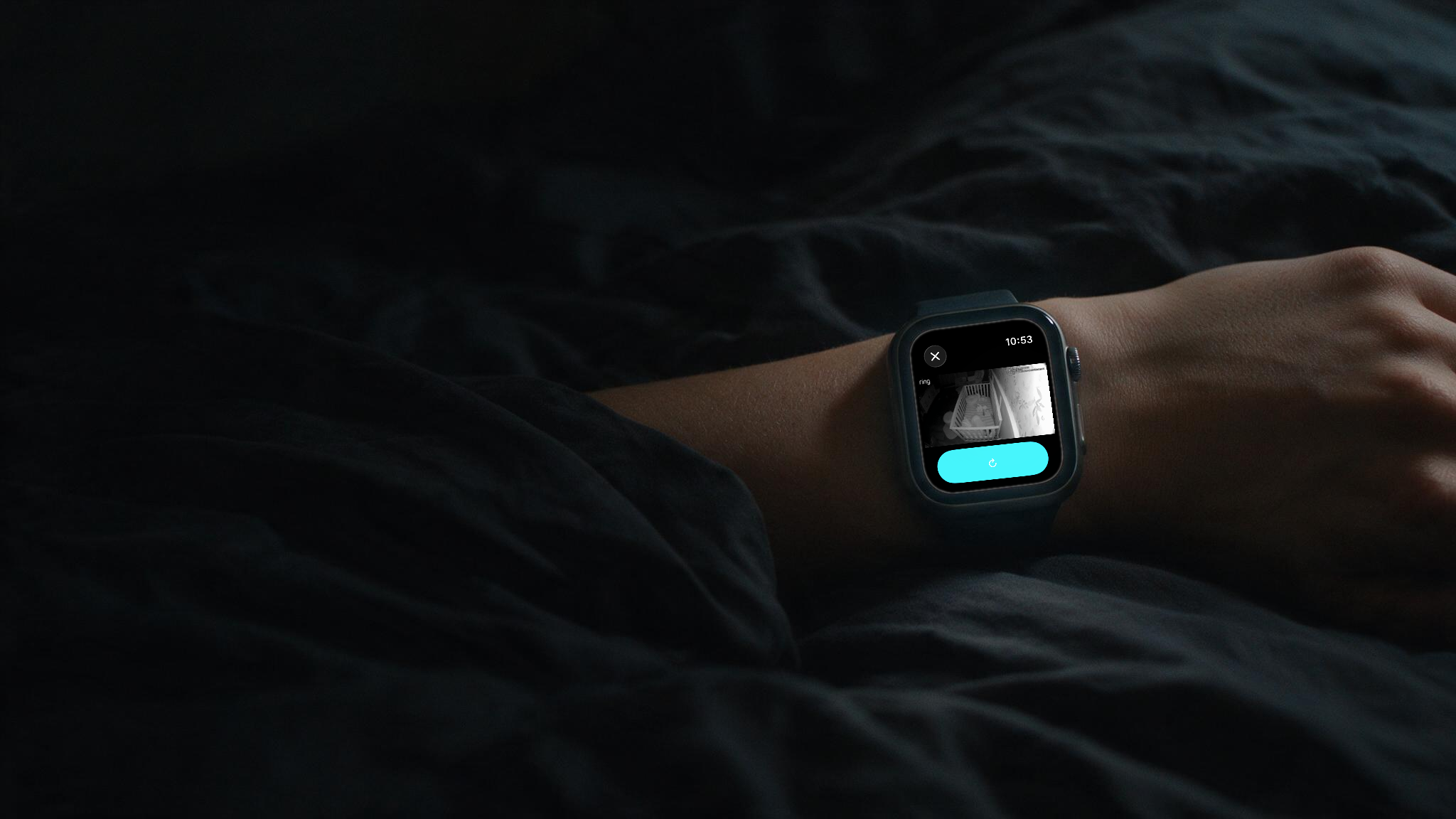 A wrist resting on dark bedding at night. The smartwatch shows a night-vision view of a sleeping baby in a crib, sent from a linked Ring camera.